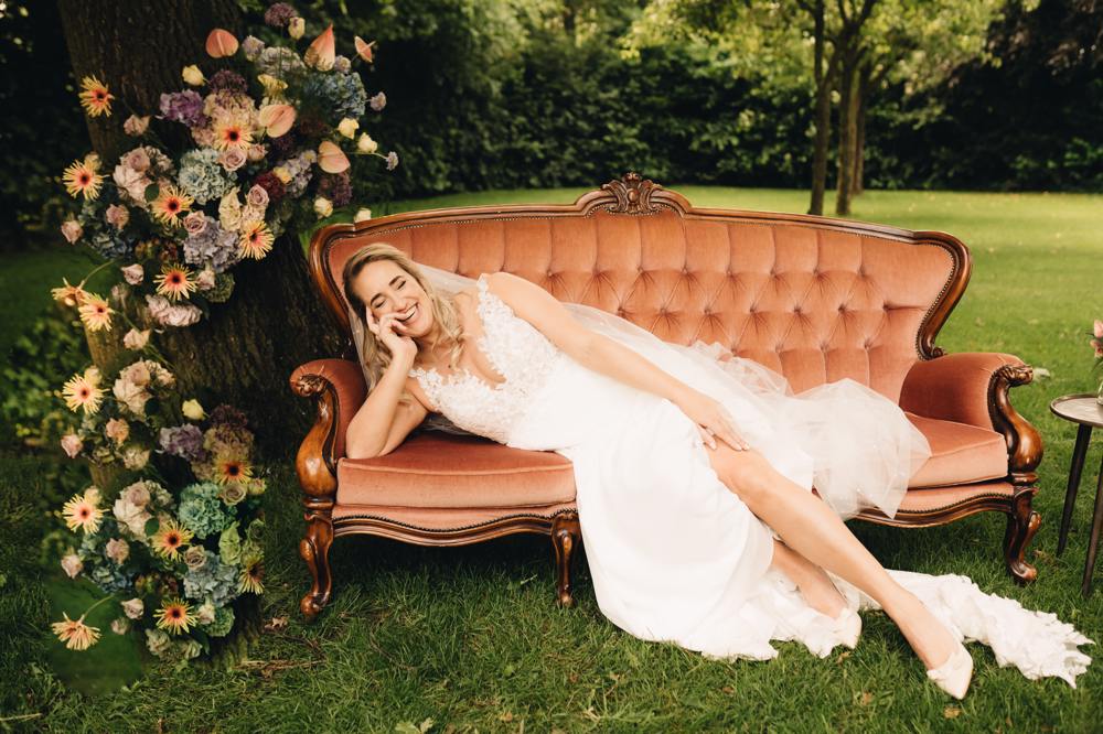 real-bride-review-dionne-van-schaick-foto-1-bruidshuis-diana – bruid lachend op vintage bank bij bloemen, tuinsetting.jpg